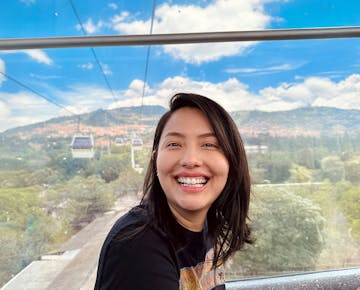 A smiling woman sitting inside a cable car with panoramic windows, with a scenic view of green hills, trees, and a bright blue sky with scattered clouds in the background.