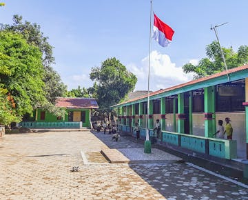 A bright school courtyard in Lombok with green and yellow buildings, a red and white Indonesian flag flying high.
