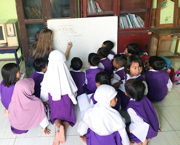 A volunteer teacher sits on the floor, drawing on a whiteboard while a group of young students in purple and white uniforms attentively watch and learn.