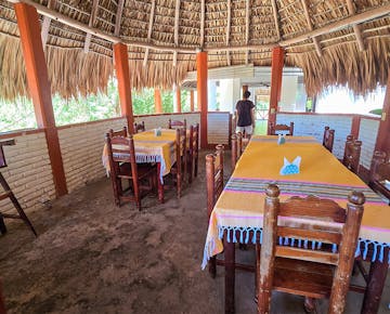 An open-air dining area in a thatched-roof restaurant, used for volunteer meals. featuring wooden tables covered with tablecloths.