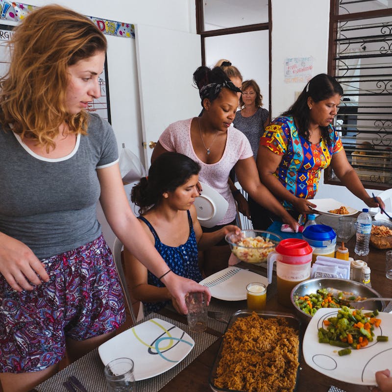 A diverse group of people gather around a table in a communal dining setting, serving and sharing a meal together. The table is filled with plates of rice, vegetables, and other home-cooked dishes.