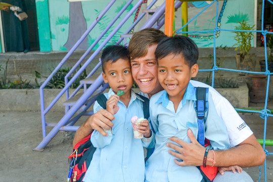 A young volunteer smiles with two local children in matching light blue school uniforms. They are outside a colorful childcare playground in Senggigi, Lombok - with one child holding a treat.